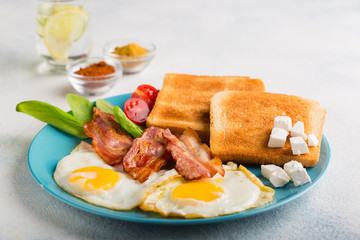 Two fried eggs with slices of bacon, toast, slices of cheese, tomatoes, spinach and a glass of mineral water on a blue plate. English breakfast. with copy space for your text