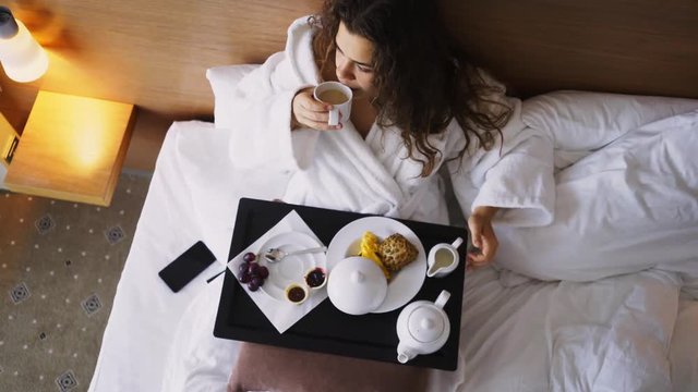 Girl enjoying morning in modern hotel apartment. Overhead view woman in white bathrobe lying in bed and have breakfast on small table-tray, pouring milk to cup of coffee. 
