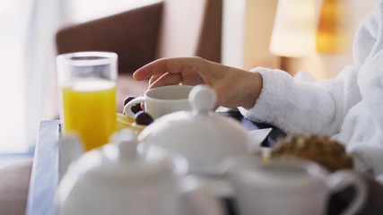 Girl enjoying morning in light hotel apartment with full length windows. Woman in white bathrobe having breakfast in bed, detail view tray full of food and female hand taking cup of tea  - Powered by Adobe