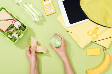 Fototapeta na wymiar cropped view of woman hands with sandwich and glass of water near lunch box, backpack, digital tablet, bottle of water and stationery