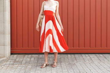 Details of stylish woman's summer outfit. Red and white midi silk dress, wicker straw bag and gold...