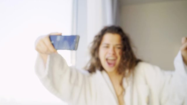 Woman having fun in apartment. Blurred female in white bathrobe taking selfie near window in backlit. In focus hand with smartphone and happy girl making face on camera. 
