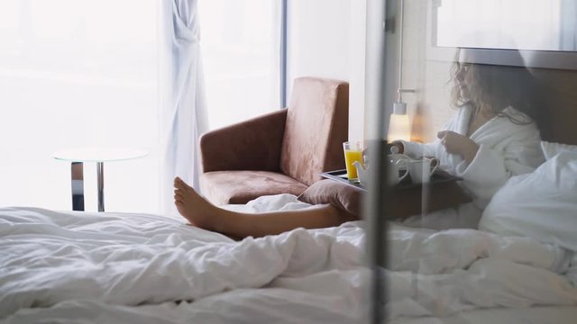 Girl in light hotel apartment with full length windows. Side view woman in white bathrobe lying in bed and have breakfast on small table-tray, enjoying coffee at morning time