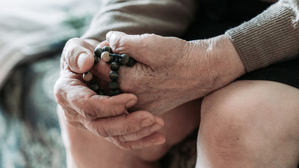 Praying hands of an old woman holding rosary beads. Coronavirus risk group. COVID virus infection.