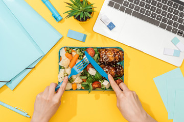 cropped view of woman holding fork over lunch box with food near laptop and office supplies