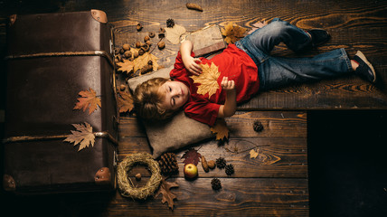 Blonde little boy resting with leaf on stomach lies on wooden floor in autumn leaves. The biggest...