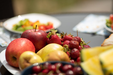 Closeup of tasty red cherries standing on plate on table. Fresh fruits and vegetables served for picnic in summer. Yellow apples and juicy melon lying at background. Concept of food and vitamins.