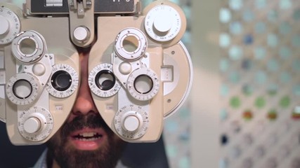 Close up shot of optometrist in white coat changing lenses on phoropter instrument and talking to male patient having eye exam . - Powered by Adobe