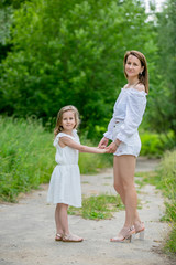 Naklejka na ściany i meble Beautiful young mother and her little daughter in white dress having fun in a picnic. They stand on a road in the park, holding hands, and look into the camera. Maternal care and love. Vertical photo