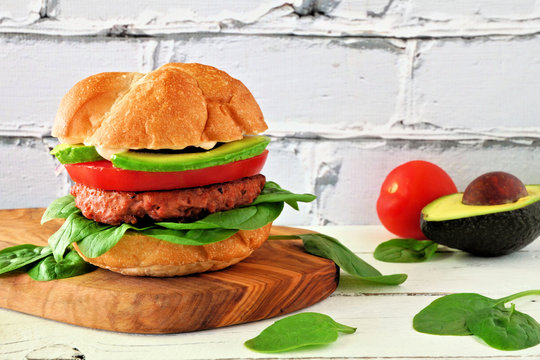 Plant based meatless burger with avocado, tomato and spinach on a wood serving board against a white brick background