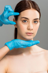 cropped view of plastic surgeon in latex gloves near attractive woman with marks on face isolated on grey