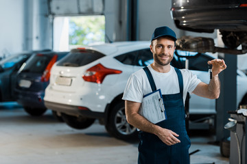 happy bearded car mechanic looking at camera and holding clipboard and key near cars