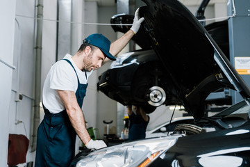 selective focus of bearded car mechanic in gloves looking at car engine