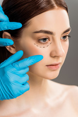 cropped view of plastic surgeon in latex gloves near woman with marks on face isolated on grey