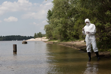 full length view of water inspector in protective costume, latex gloves and respirator holding flask with water sample at river
