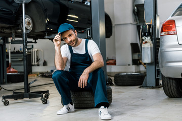 Fototapeta na wymiar happy bearded man touching cap and sitting on car tire
