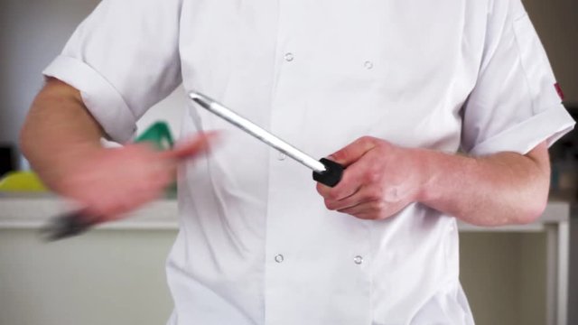 professional male chef sharpening a knife with a steel in his chef whites ready to cook a gourmet meal, then checks with his thumb to make sure it's sharp.