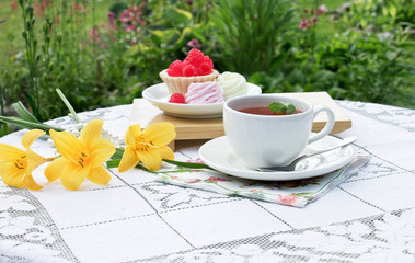 On a dacha table with an openwork tablecloth there is a cup of tea,dessert and flowers