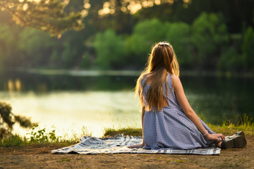 Girl blonde in a light dress sitting on a blanket and looking into the distance at sunset. Girl at sunset. Horizontal photography