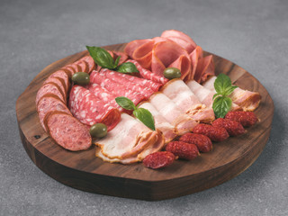 Antipasto set platter on wooden plate over gray stone. Cold smoked meat plate with sausage,sliced ham,prosciutto, bacon, olives and basil. Appetizer on wooden tray cut tree sawed imitation. Copy space