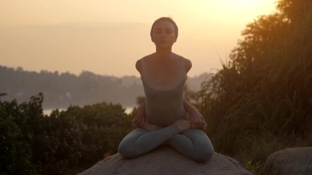 yoga teacher shows baddha padmasana with forward bending on huge rock against tropical trees low angle shot slow motion. Concept fitness yoga wellness lifestyle