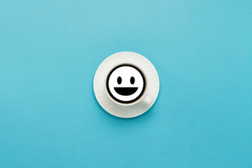 White cup on a saucer, black coffee, blue background. Sign face with a smile. Flat lay, top view