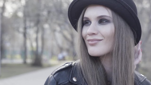 Smiling attractive stylish girl with smoky eyeshadow wearing black hat and leather jacket. Action. Young adorable brunette woman with straight hair posing outdoors, looking aside.