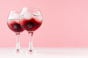 Sweet red alcoholic cocktails with ice cubes, blueberry in two goblets in soft light pink bar interior, copy space.