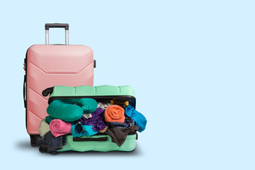 Opened plastic suitcase with wheels, crowded with things and a smaller suitcase standing next to a blue background. Travel concept, vacation trip, visit to relatives