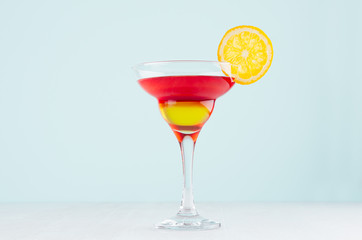 Summer fresh alcohol cocktail  with red and yellow liquor, orange slice on soft light white and pastel green background.