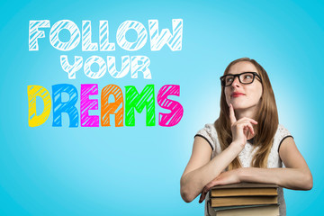 Young girl student with glasses leaning on a pile of books and looking up with a pensive face on a blue background with text Follow your dreams