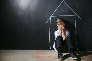 Homeless little boy sitting on floor near dark wall