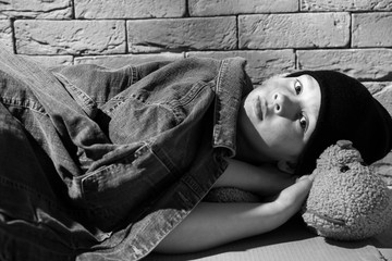 Homeless little boy with teddy bear lying on floor near brick wall