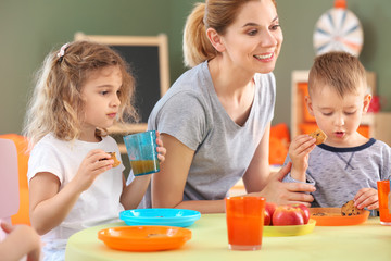 Nursery teacher with cute little children during lunch in kindergarten