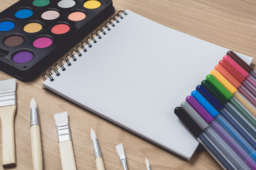 Notepad or notebook with Many colorful pens, paintbrush and Watercolor palette on brown wood table.using for Arts and Education background