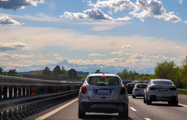 View of cars on highway in traffic jam with the mountains of the Alps in the background and lens flare at sunset, Piedmont, Italy