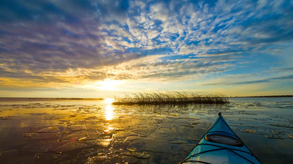 View from bow (prow) of blue kayak on lake with ice floes in winter or early spring at buatiful sunset. Winter kayaking