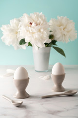 Obraz na płótnie Canvas Boiled eggs in ceramic egg cups with ceramic spoons on marble table top decorated with beautiful white peony. Breakfast concept. Close-up.