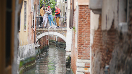Four young friends standing on the bridge between two buildings above the water channel - two women hug - Venice, Italy
