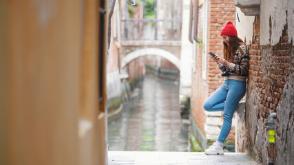 A young woman standing near the water channel and searching for something in her phone - Venice, Italy