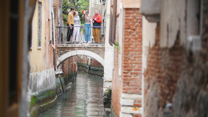 Four young friends standing on the bridge between two buildings above the water channel - Venice, Italy