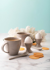 Boiled eggs in ceramic egg cups, two cups of coffee with ceramic spoons on background of beautiful white peony. Breakfast concept. Copy space.