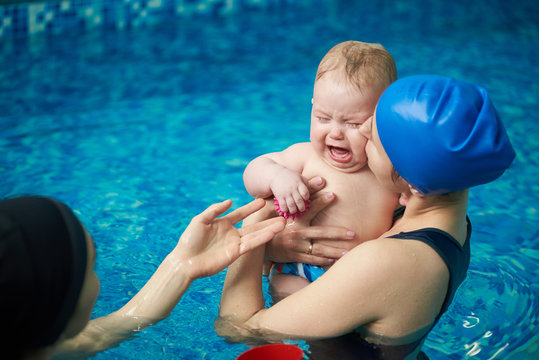 Little blond baby boy in diaper crying, afraid of water and swimming. Mother calming baby, holding on hand and kissing her baby. Women accustoming small child to water and teaching swimming in pool