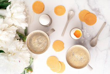 Soft-boiled egg with liquide orange yolk in ceramic egg cups, two cups of coffee with ceramic spoons on background of beautiful white peony. Breakfast concept. Top view.