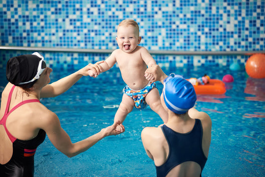 Women holding toddler lifting high up while having fun in water. Baby in swimming nappies rejoicing and showing tongue. Activities in swimming pool. Doing stretching exercising over water for infant