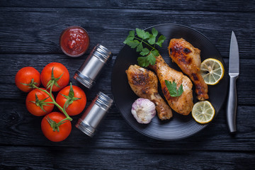 Grilled chicken legs with tomatoes, lemon, parsley and ketchup on a black plate. Top view