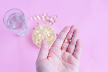 one capsule of omega three on the female palm on a pink background with a glass of water and fish oil top view