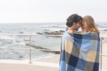 Fototapeta premium Caucasian couple wrapped in blanket standing at promenade near beach 