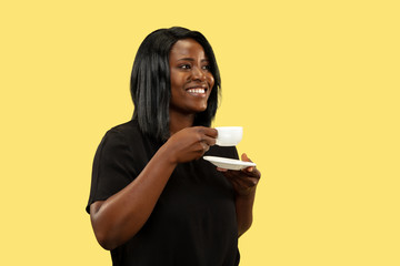 Young african-american woman isolated on yellow studio background, facial expression. Beautiful female half-length portrait. Concept of human emotions, facial expression. Enjoying of drinking coffee.