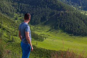 Guy in a green t-shirt is standing  on the edge of the cliff in rear of him is a colorful view of mountains  and green trees. Traveler complete the route